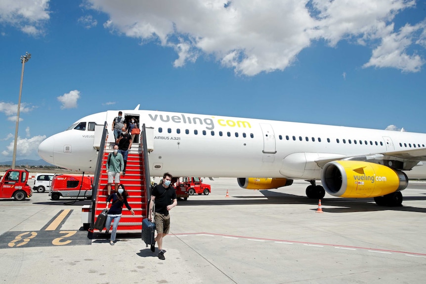 On a bright, but cloudy day, you view passengers taking exit stairs on a small white passenger jet with yellow engines.