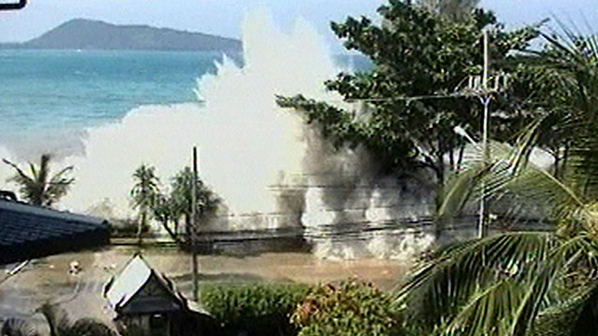 A video still of a tsunami wave coming over a wall and sending up a big spray of water. The blue ocean is in the background