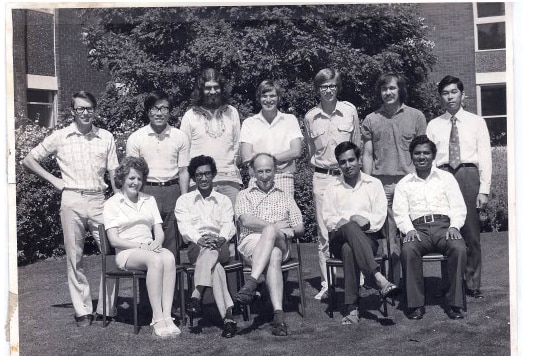 John Bockris, bald and wearing a white shirt, sits in the middle of a group of younger researchers in a black and white photo.