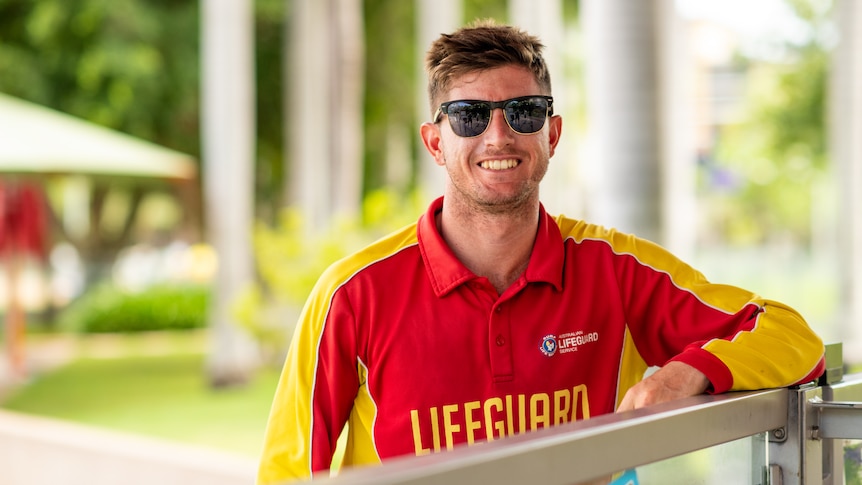 A pool lifeguard wearing red and yellow uniform wearing glasses smiles at the camera.