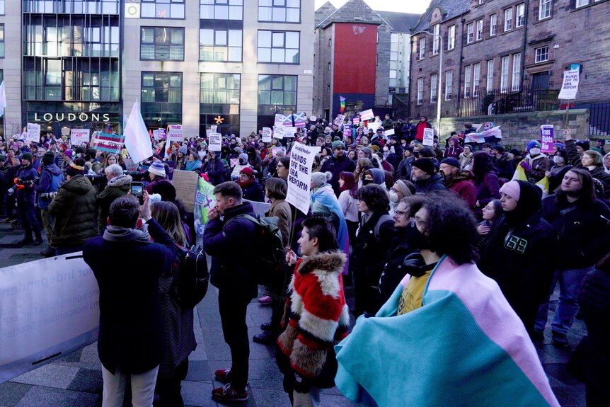 Hundreds of people gather in a paved courtyard with placards and flags striped pink, white and blue