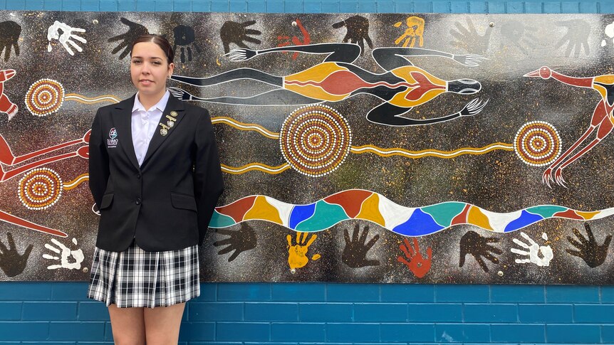 Music artist Llaney Webb stands in front of a brick wall with an indigenous design