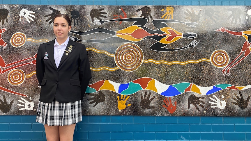 Music artist Llaney Webb stands in front of a brick wall with an indigenous design