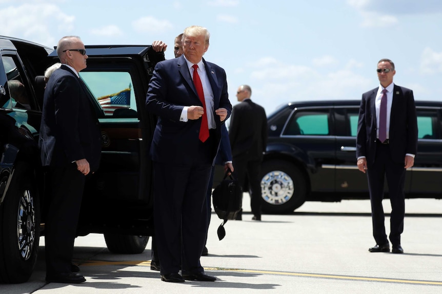 Donald Trump stands flanked by presidential vehicles and secret service officers in dark suits.