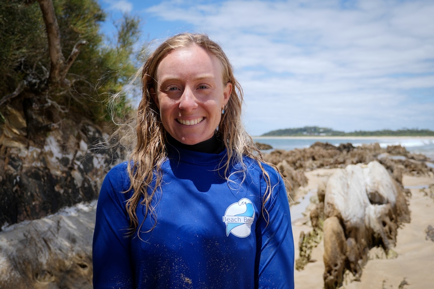 Una mujer en traje de neopreno sonríe a la lente de la cámara con una playa al fondo. 