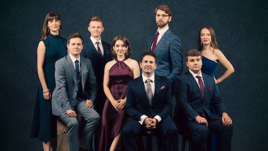 Vocal ensemble Voces 8 posing for a photo dressed in suits and formal dresses against a dark background