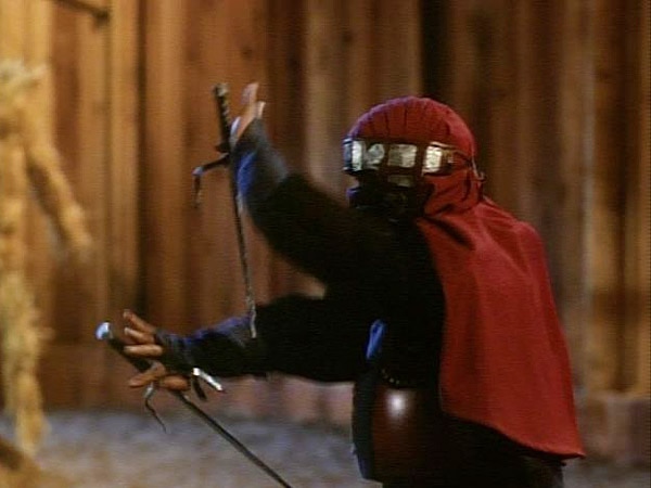 Man in masked cape holding pair of sais in fight scene of a movie.