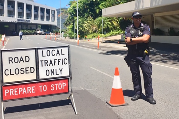 A police officer stands next to a traffic cone and sign that says ROAD CLOSED. He has his arms crossed over his chest.