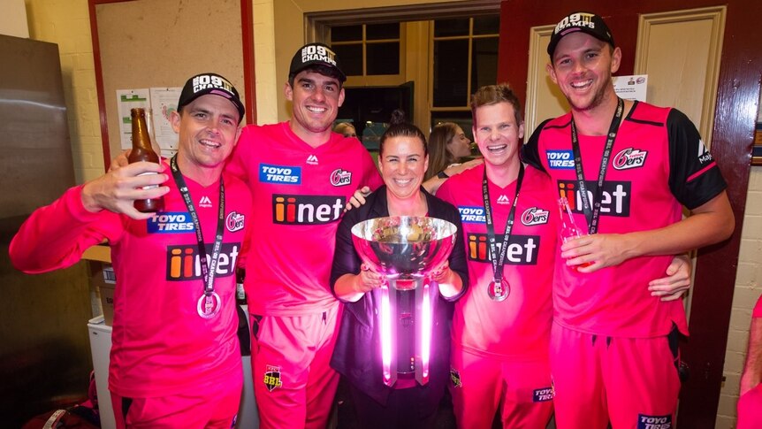 Steve O'Keefe, Moises Henriques, Jodie Hawkins, Steve Smith and Josh Hazlewood stand, wearing pink, holding a trophy