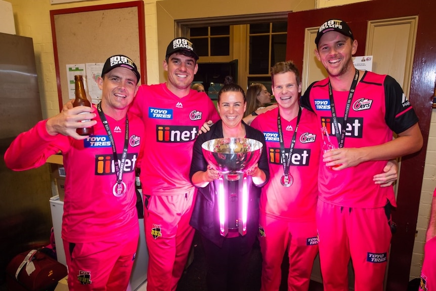 Steve O'Keefe, Moises Henriques, Jodie Hawkins, Steve Smith and Josh Hazlewood stand, wearing pink, holding a trophy