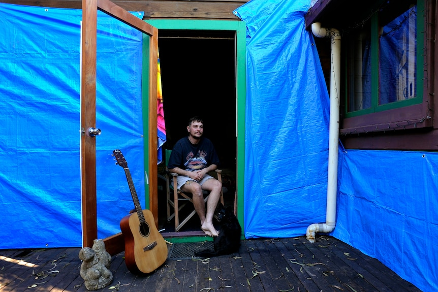 A man sits with a dog in in the doorway of a damaged house draped in blue plastic.