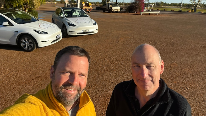 Two men stand together. Behind them are two cars, a blue sky and red dirt.