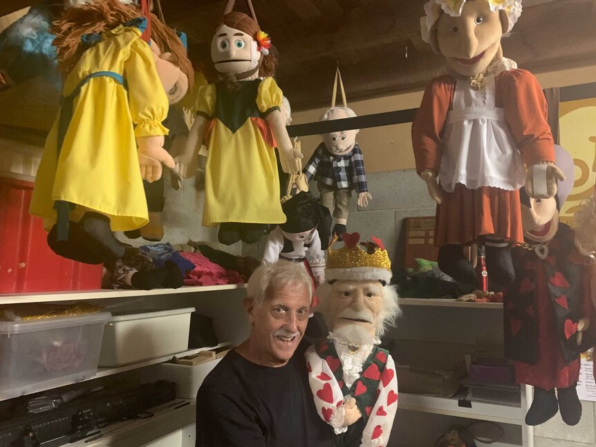 A man with white hair and a moustache holds a puppet of a king as various puppets hang from the ceiling