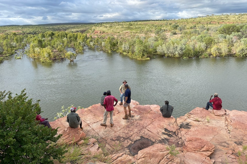 A group of rangers stand and sit on a rocky outcrop, overlooking a river with wetlands in the background and a cloudy sky.