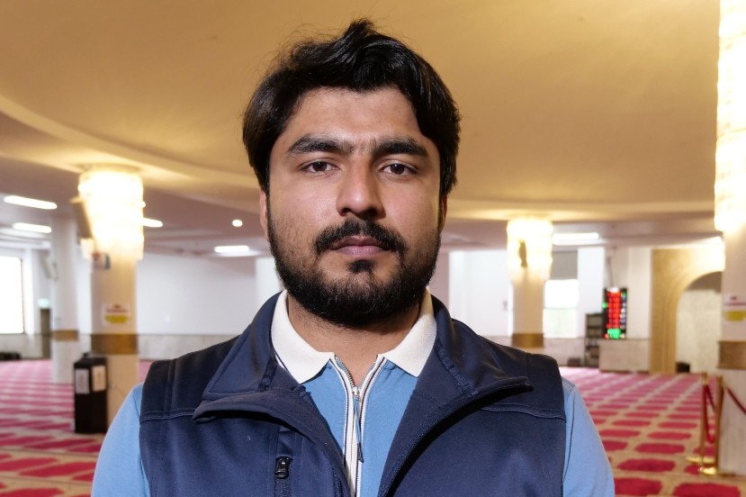 a man with a beard and a moustache looks at the camera as e attends a prayer service for his dead cousin