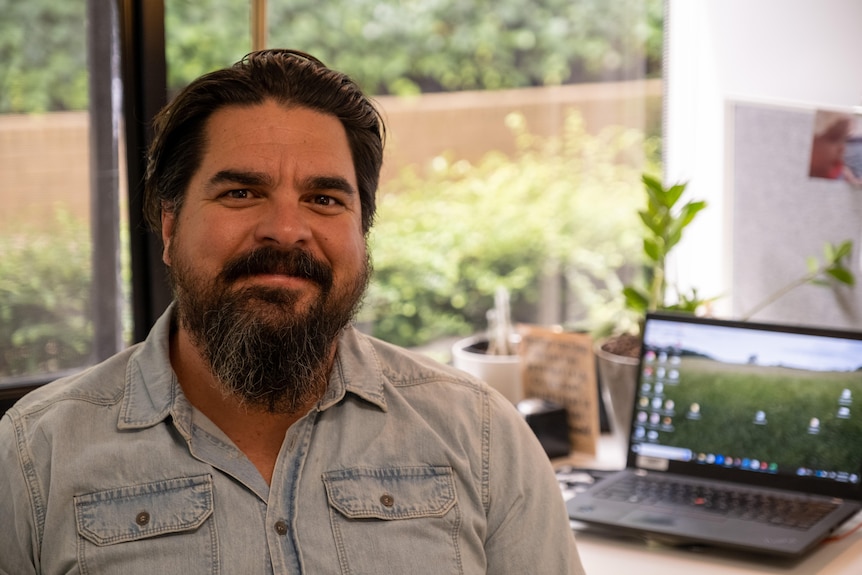 A man in his 40s with black hair and a beard, smiling in front of a laptop. 