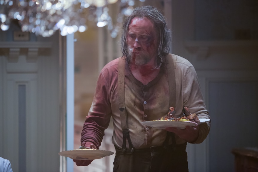 A battered-looking Nicolas Cage with long greying hair and a blood-soaked shirt holds a dinner plate in each hand