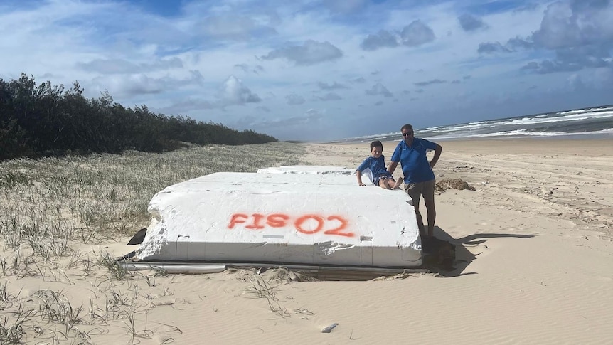 Man and a child lean against a pontoon washed onto a beach