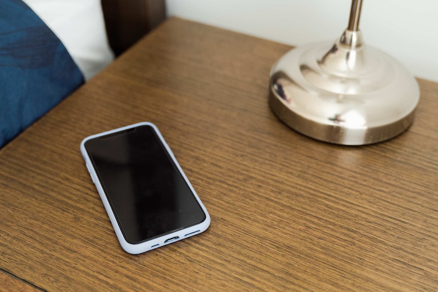 A mobile phone lies on a bedside table near a lamp.