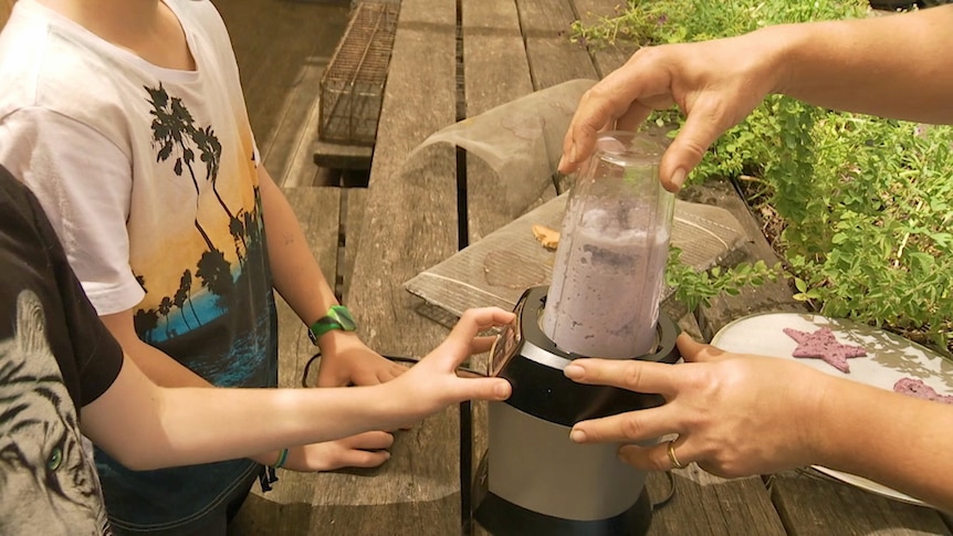 Two children and woman are blending a purple mix in a blender with cut-out purple shapes on a plate nearby