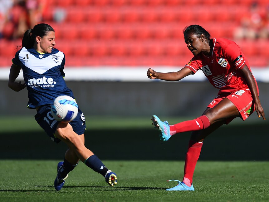 A soccer player wearing red kicks the ball at a player wearing blue