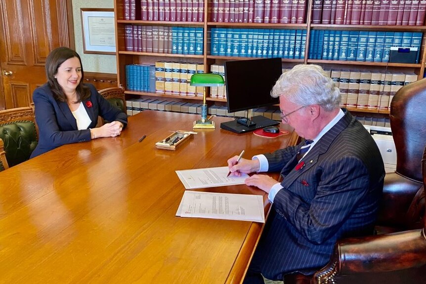 The Queensland Governor signs a document while the Queensland premier looks on.