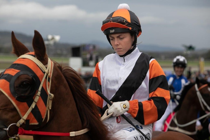 a small woman jockey wearing white black and orange sitting on the back of a race horse after competing.