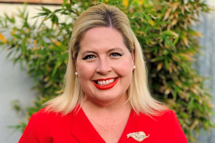Katrine Hildyard stands smiling in a red shirt with a gold broach on the collar