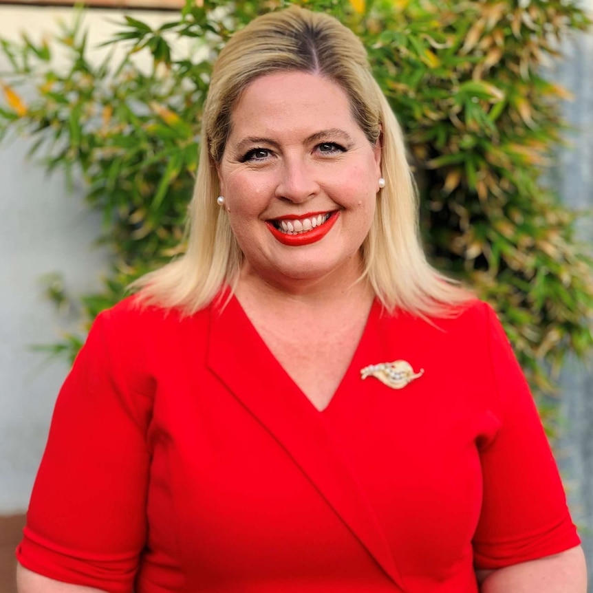 Katrine Hildyard stands smiling in a red shirt with a gold broach on the collar