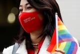 A person in a red COVID mask holds a rainbow flag up near her face. 