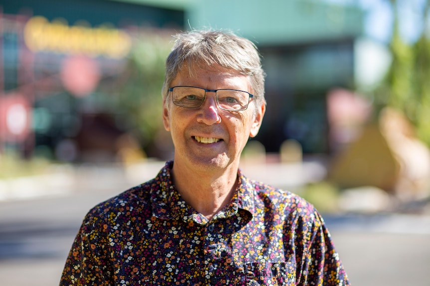 A man wearing glasses and a floral shirt smiles at the camera outside in the sun.