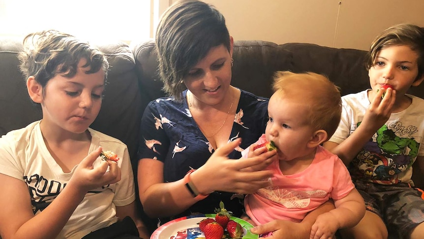 Angela Stevenson sits on a couch in her home eating strawberries with her three children.