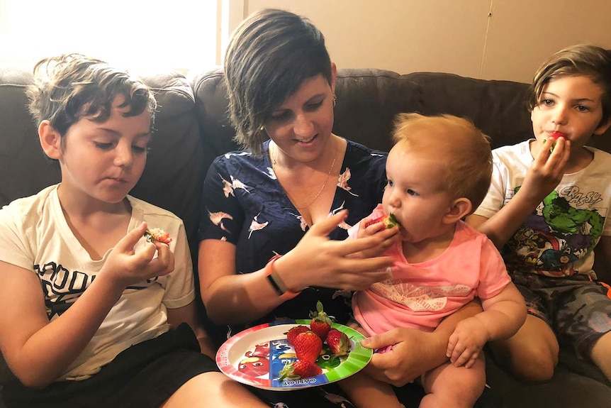 Angela Stevenson sits on a couch in her home eating strawberries with her three children.