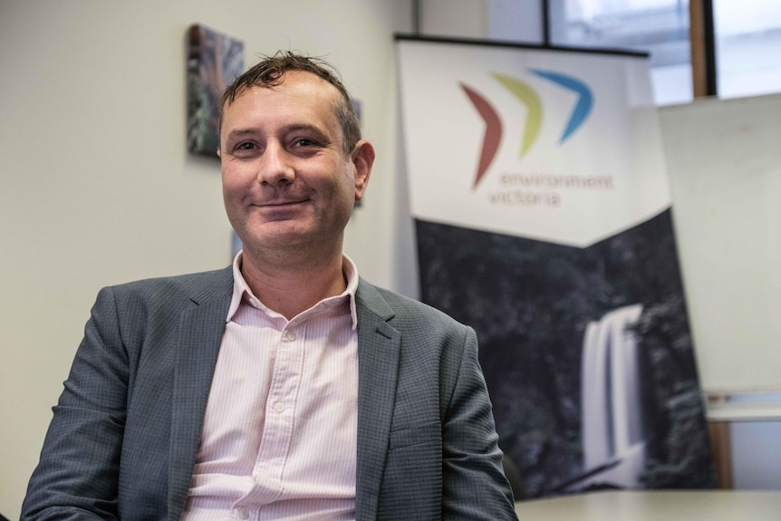 Jono La Nauze, wearing a pink button-up shirt and grey jacket, sits in an Environment Victoria office.