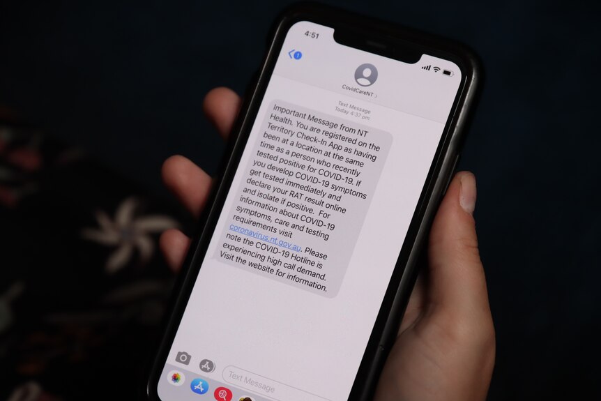 A person is holding a phone with a text message from COVID Care, urging them to watch for symptoms and get a RAT if required.