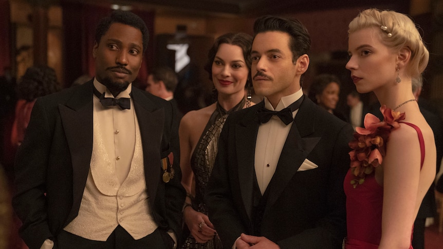 One black man, a moustached white man, and two white women dressed in fancy 1930's ballroom attire in a fancy function. 