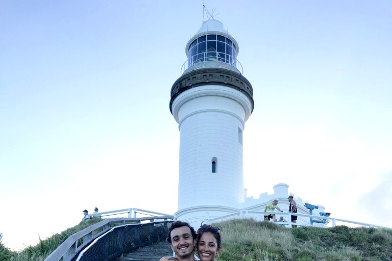 A tall tan man smiles for a photo with a woman's arms wrapped around him in front of an old white lighthouse.