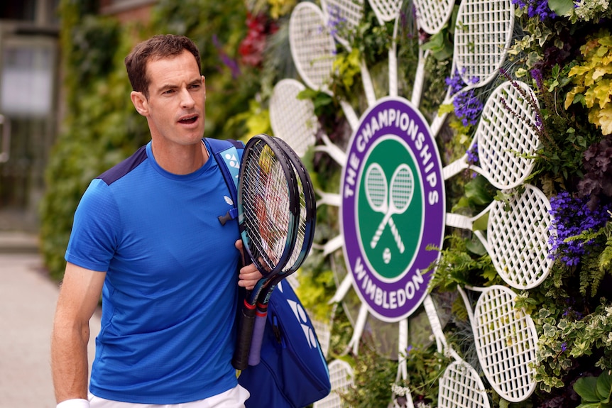 Tennis veteran Andy Murray walks past a WImbledon sign carrying a bag over his shoulder and a couple of tennis racquets.