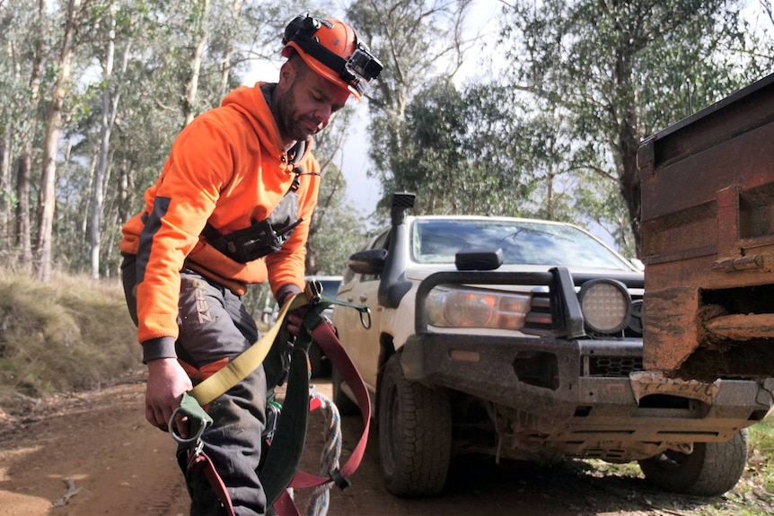 A man stands on a dirt road in a forest and puts his leg through a safety harness.