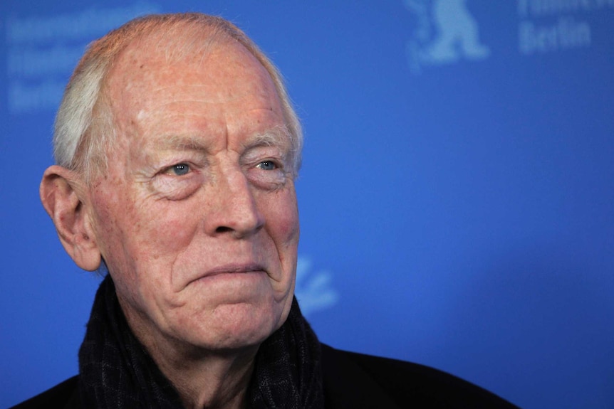 Max von Sydow looks to the right and smiles. He stands in front of a blue background.