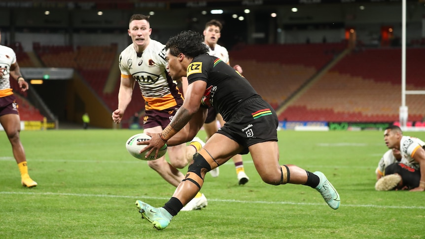 A Polynesian rugby player in a brown jersey scores a try as players in white and yellow look on. 