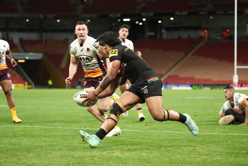 A Polynesian rugby player in a brown jersey scores a try as players in white and yellow look on. 