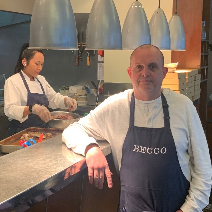 Simon Hartley stands in a 'Becco' branded apron in front of the kitchen of his restaurant.