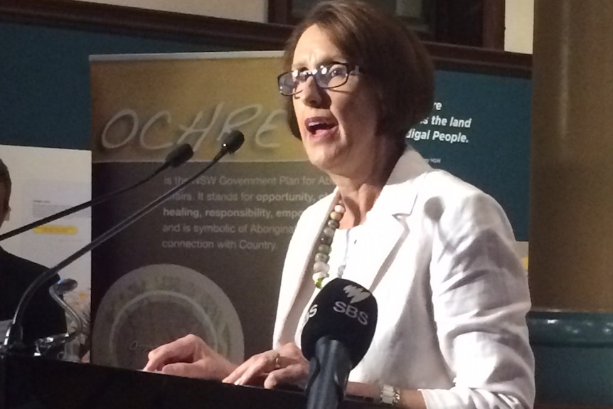 A dark haired, bespectacled woman in a white suit speaks at a lectern.