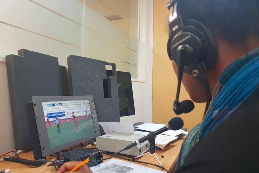 A woman with headphones looks at a TV screen with female players on a pitch