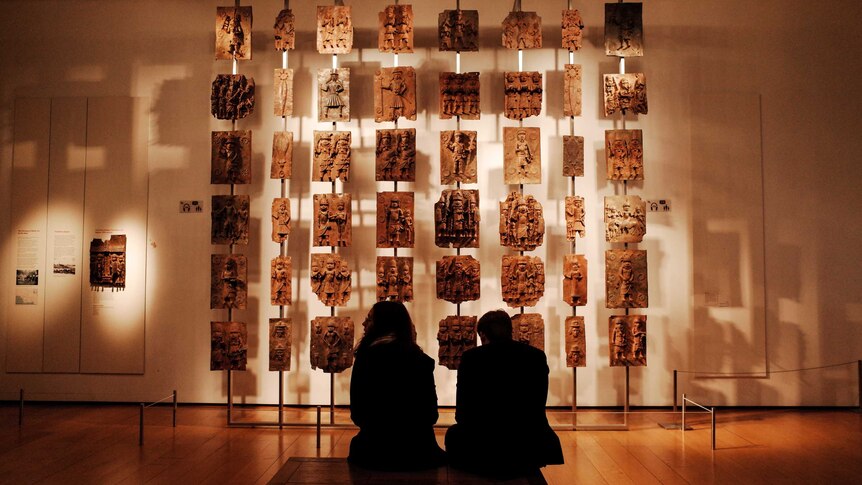 Two museum visitors sit looking at a number of sculptured plaques arranged hanging in front of a wall.