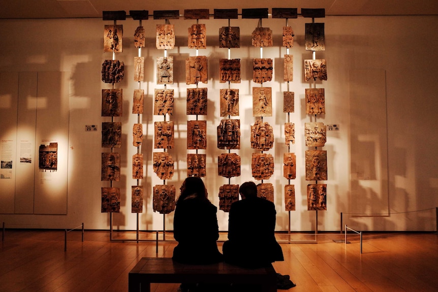 Two museum visitors sit looking at a number of sculptured plaques arranged hanging in front of a wall.