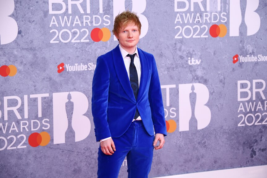 A red haired man in a bright blue suit stands awkwardly on the red carpet