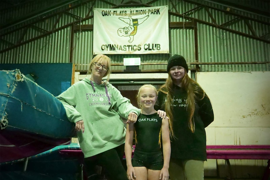 Kim Lacey, Paytience Hesse and Jess Kane stand below a sign saying Oak Flats Albion Park gymnastics club.