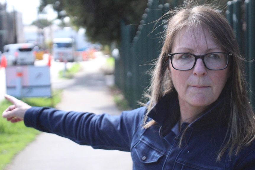 A woman wearing a blue top, glasses points in a direction away from the camera. Looks serious.